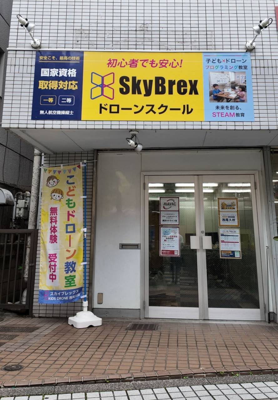 スカイブレックス KIDS DRONE 西尾久校の外観。黄色いSkyBrexの看板と「こどもドローン教室 無料体験受付中」ののぼりが目印