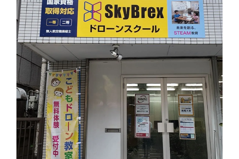 スカイブレックス KIDS DRONE 西尾久校の外観。黄色いSkyBrexの看板と「こどもドローン教室 無料体験受付中」ののぼりが目印