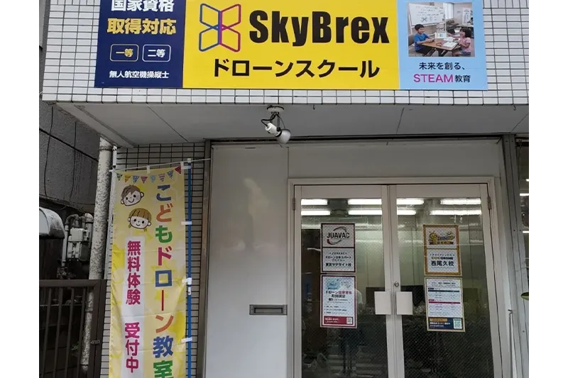 スカイブレックス KIDS DRONE 西尾久校の外観。黄色い看板とオレンジののぼりが目印