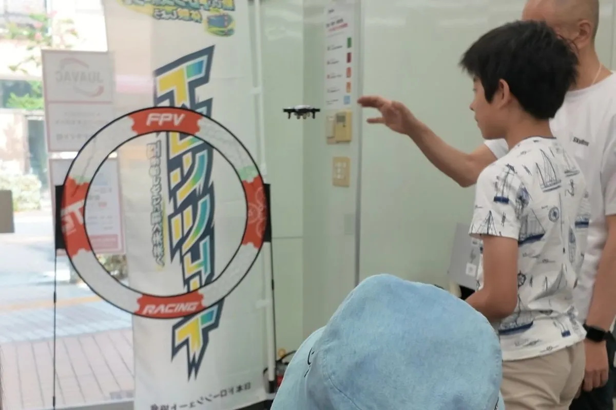 安全ネットを張った屋内スペースで、小学生が見守られながらドローンを飛ばしている様子（東京都荒川区西尾久）